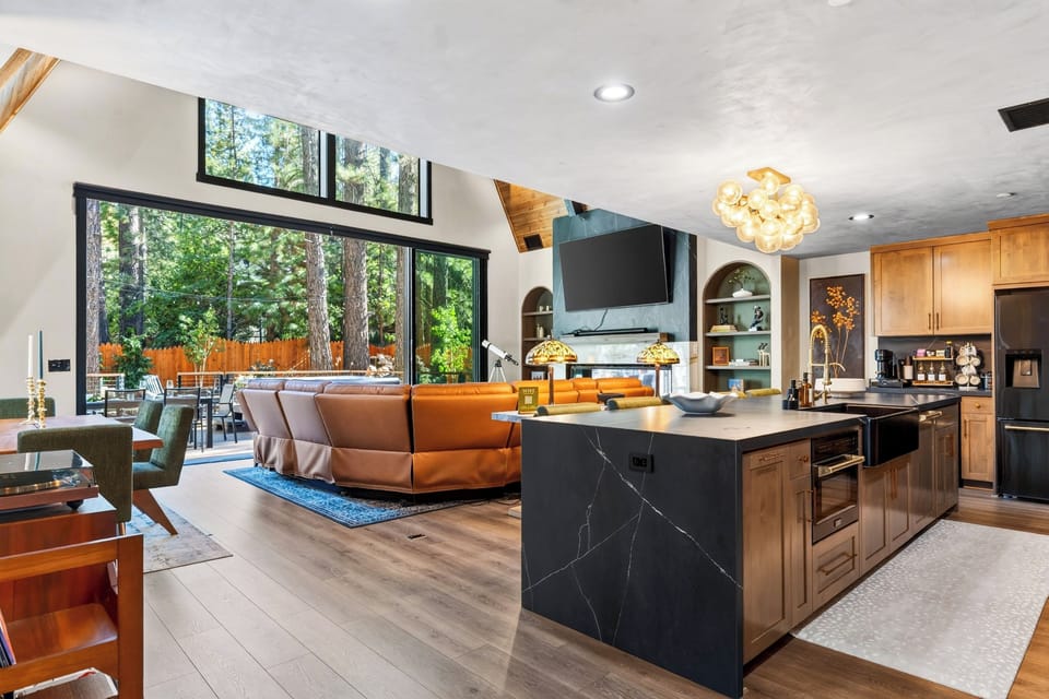 The open-concept great room brings the kitchen and lounge together beneath soaring wood ceilings and expansive forest-facing windows.