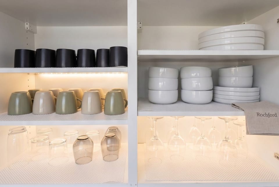 The kitchen cupboard holds crockery and glassware under warm shelf lighting.