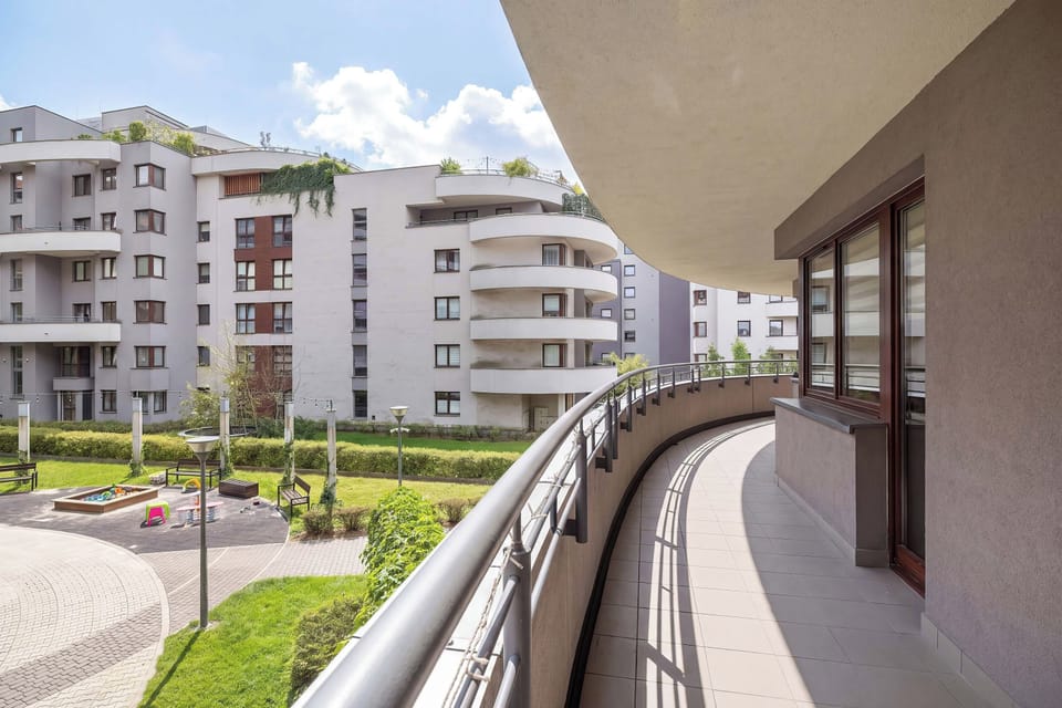 A curved balcony with outdoor furniture overlooking the surrounding apartment buildings and greenery. The elevated position provides an open view and a pleasant setting for relaxing outdoors.