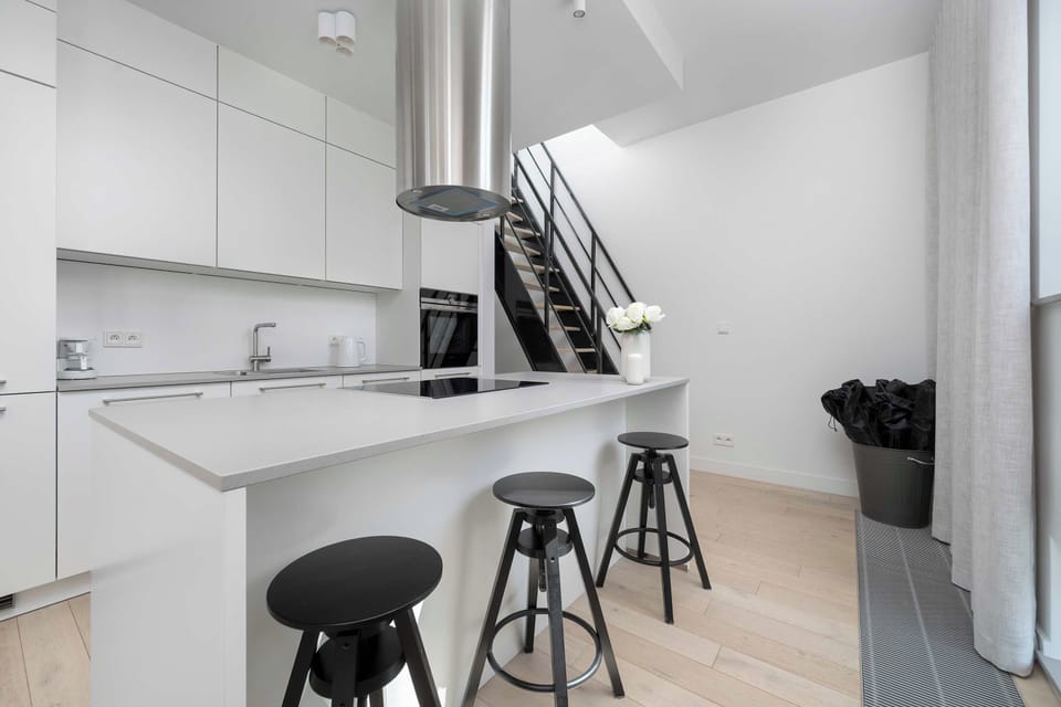 A modern kitchen island with black barstools sits adjacent to a sleek staircase. Stainless steel and white finishes reflect the home's contemporary architectural flair.