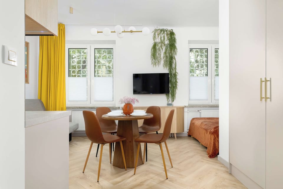 Natural light fills the studio space through large windows with vibrant yellow drapes. A round dining set sits centrally on the light wood flooring.