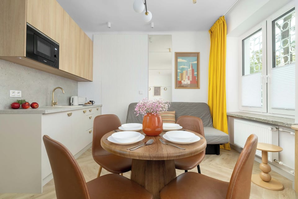 The dining area includes a round table neatly set with dinnerware and comfortable chairs. Kitchen cupboards and large windows frame the background.