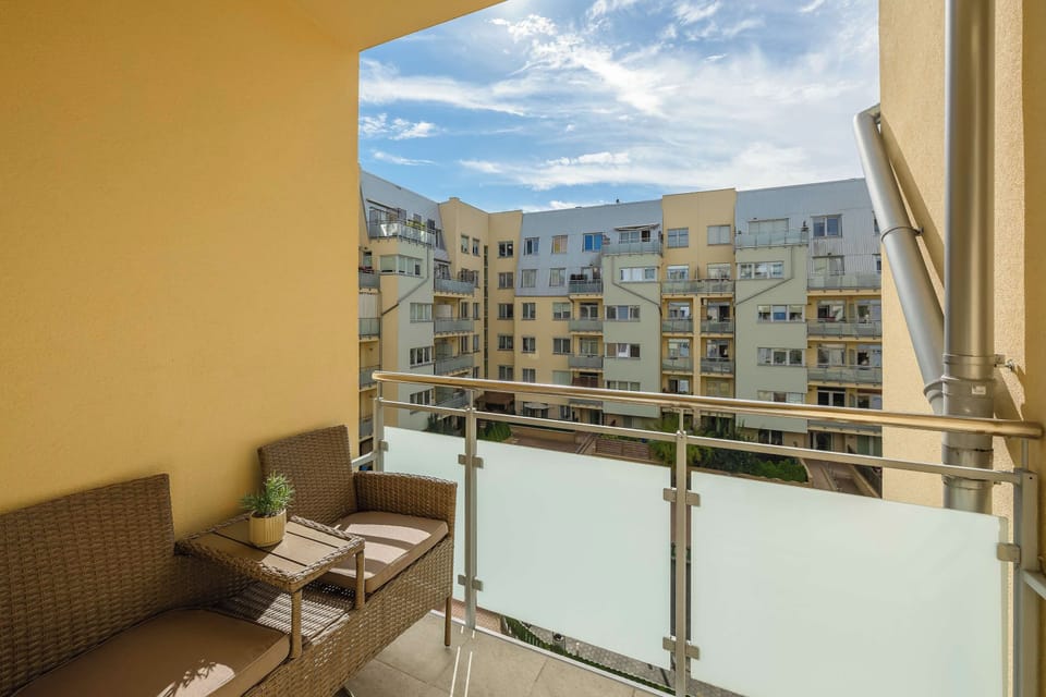 An outdoor balcony equipped with wicker seating and a small table. Glass railings offer a clear view of the surrounding residential buildings.