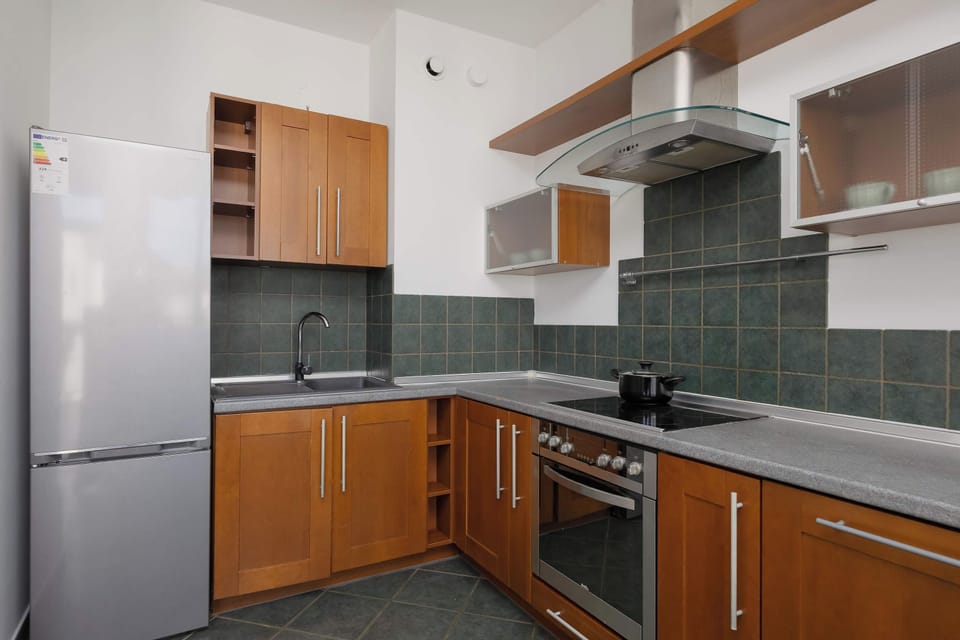 A modern kitchen displaying wooden upper and lower cabinets with a dark green backsplash. A tall stainless steel refrigerator stands at the far end.
