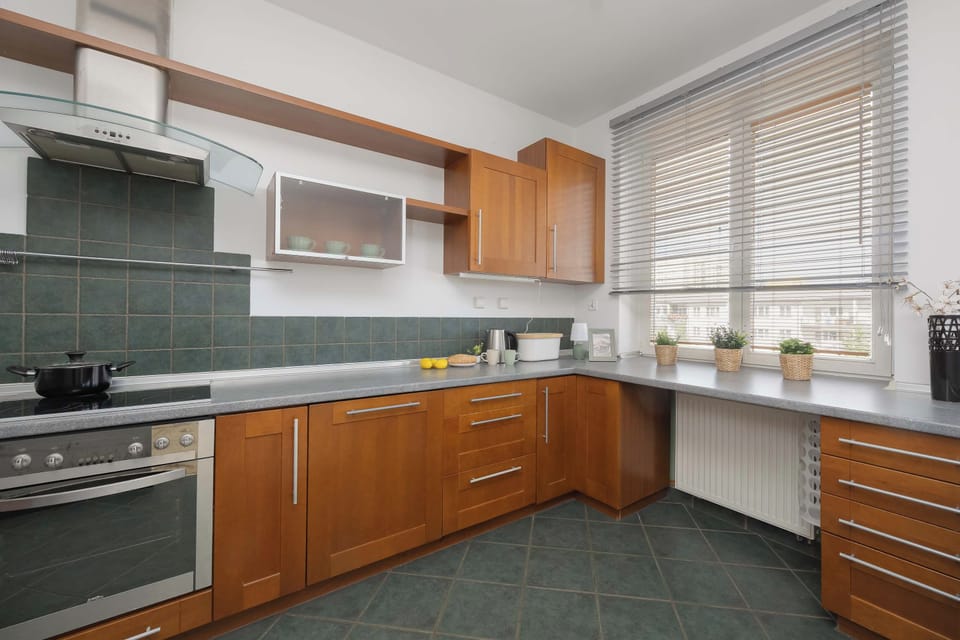 A well-lit kitchen featuring wooden cabinetry and dark countertops. Sunlight streams through a window fitted with horizontal blinds.