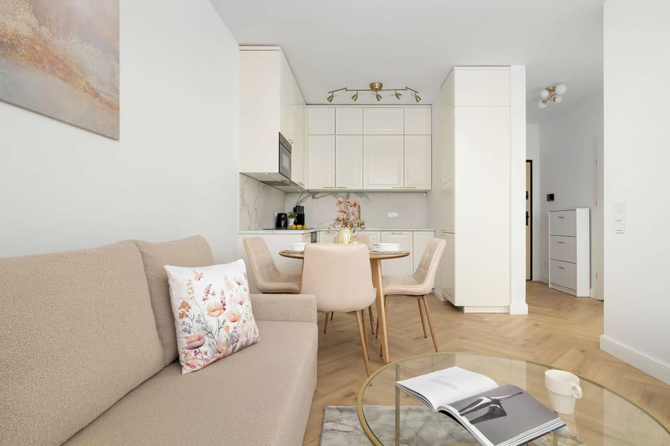 The spacious living room is bathed in natural light reflecting off the bright white walls and light wood flooring. The open layout provides clear sightlines into the adjacent kitchen and dining spaces.