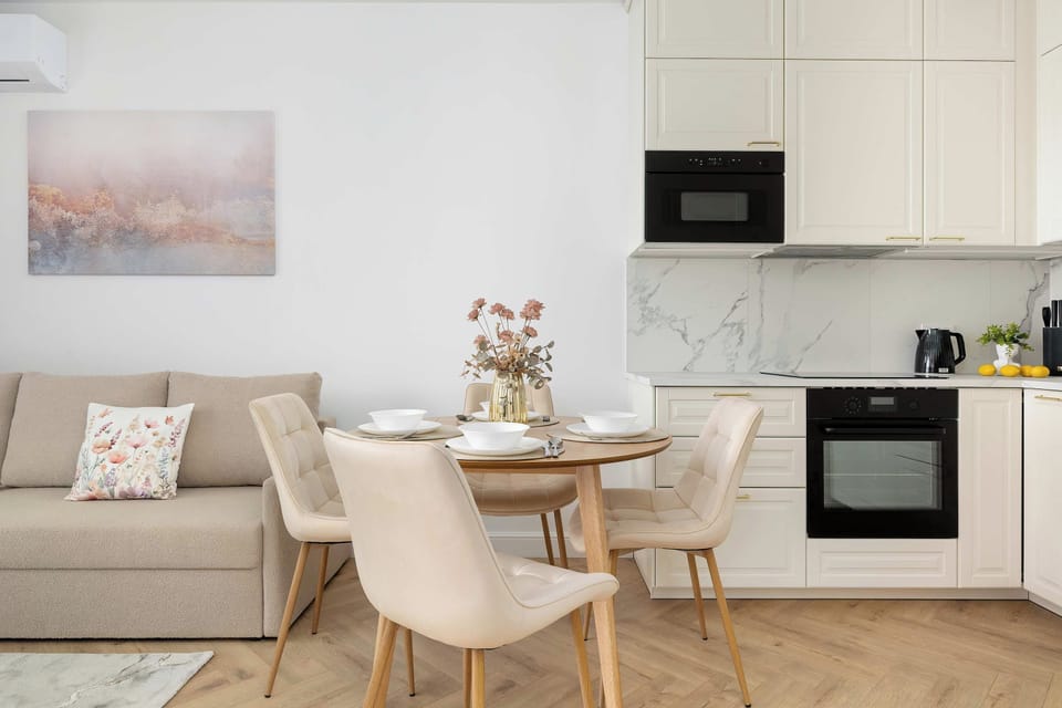 A round wooden dining table is surrounded by four stylish upholstered chairs. It sits neatly between the living room area and the modern white kitchen counter.