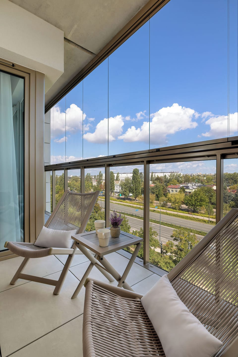 A cozy balcony space is furnished with matching woven chairs and a small table. The glass railing provides an unobstructed view of the surrounding neighborhood and greenery.