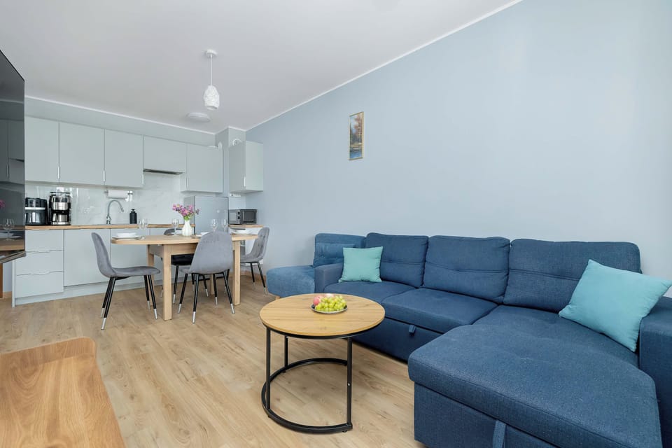 A wide view of an open-concept living space reveals a blue sectional sofa and a round dining table. The kitchen area can be seen in the background, featuring light cabinetry.