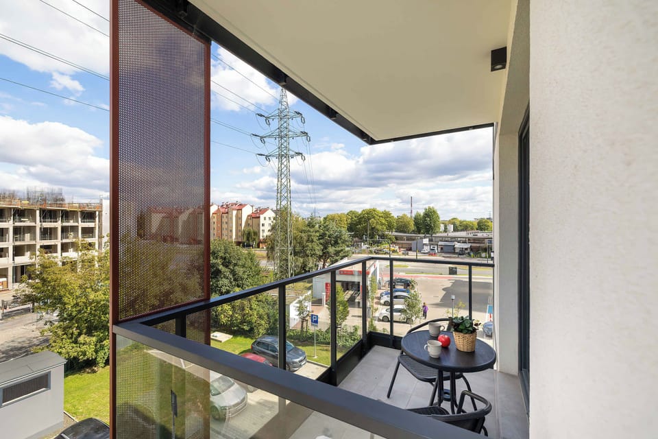 A spacious balcony with comfortable seating and a glass railing overlooking the nearby buildings and greenery. The covered outdoor area is ideal for relaxing outside.