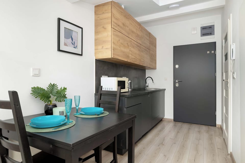 A dining area features a dark table set with blue plates and two chairs. In the background, a modern kitchen with a black countertop and wooden upper cabinets is visible. A closed dark door leads out of the apartment space.