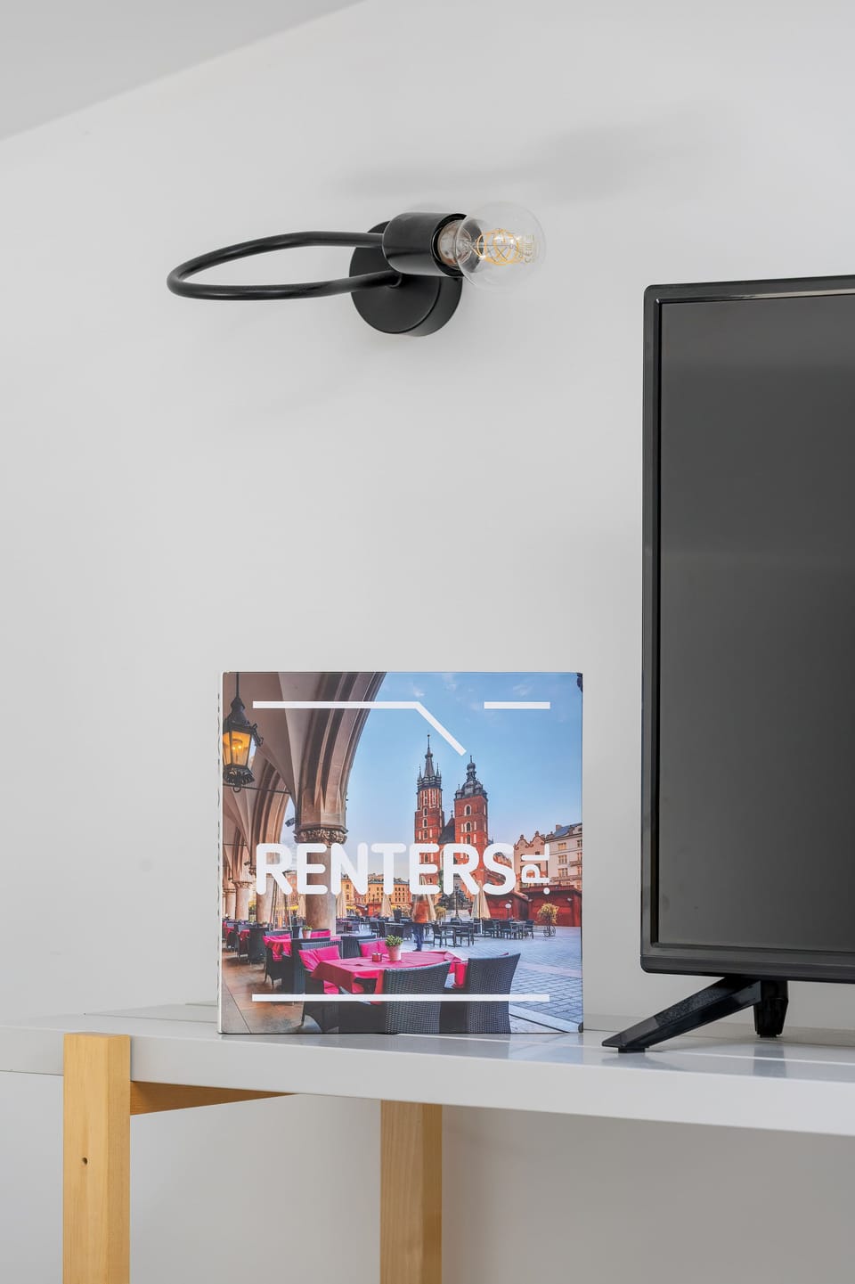 A close-up view focuses on a wall-mounted flat-screen TV in a bright room. Below the TV, a small magazine or informational booklet rests on a light wooden desk. A black wall sconce provides additional lighting above the desk area.