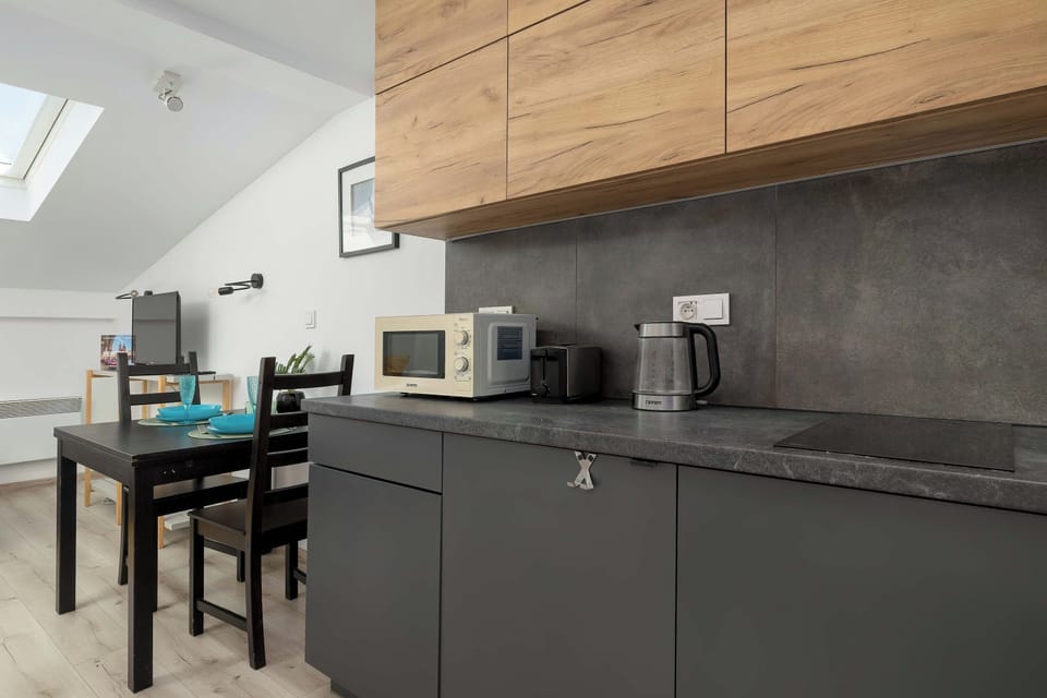 A modern kitchen area features dark gray lower cabinets and light wooden upper cabinets. A small dining table with chairs sits nearby under the slanted ceiling. A microwave and kettle are visible on the kitchen counter.