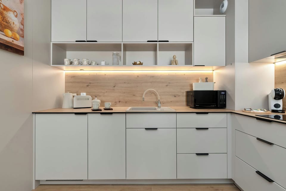 A modern kitchen with white cabinets, built-in appliances, and a light countertop. The clean, minimalist design is complemented by light wooden flooring and a bright interior.