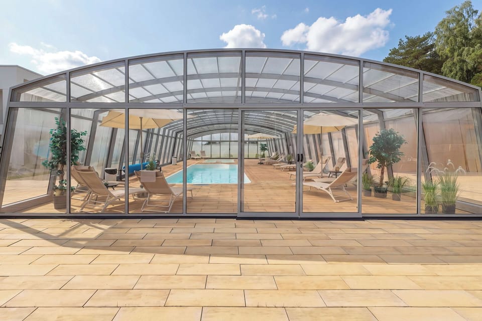 A swimming pool enclosed by a large transparent structure with an arched roof. Lounge chairs and greenery are arranged around the pool, creating a comfortable leisure area.