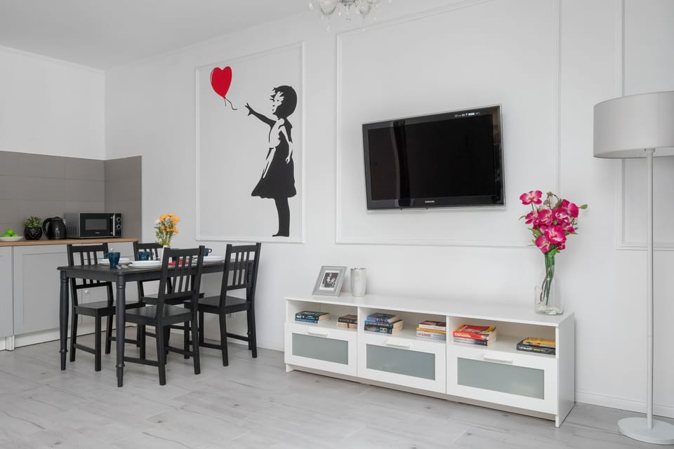 A bright dining area with a wooden table and chairs, a wall-mounted TV, and a modern kitchen in the background. White walls and light flooring create a clean and spacious atmosphere.