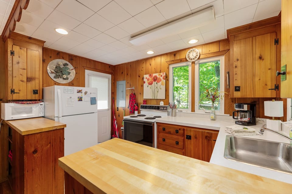 Bright and inviting bedroom with a comfortable bed, fresh linens and towels, ceiling fan, bedside lighting, and windows overlooking the peaceful wooded surroundings.