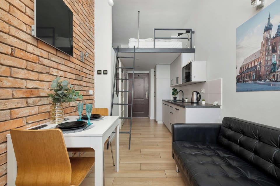 A view across the open studio showing the sofa, dining area, and kitchenette. The wooden flooring and brick accent wall create a warm contrast with the white cabinetry.