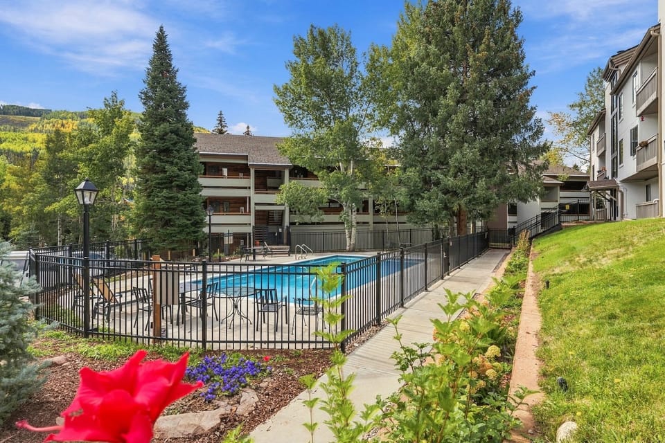 An outdoor pool surrounded by a fence, mature trees, and residential buildings.