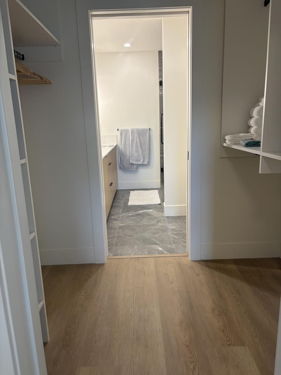 Light wood flooring leads from a walk-in closet with open shelving to a bathroom with gray tile flooring.
