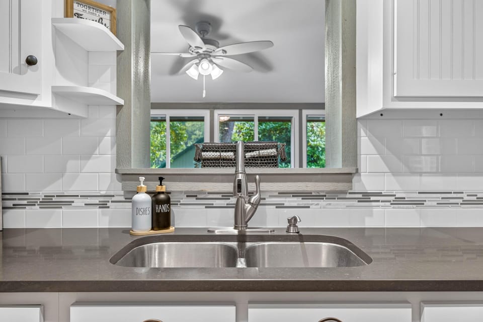 Stainless steel double sink set in dark quartz countertop with white tile backsplash and soap dispensers.