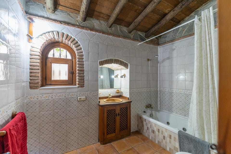 Traditional beach villa family bathroom featuring a built-in bathtub, a rustic wooden vanity sink cabinet, tiled walls, and a charming arched brick window.