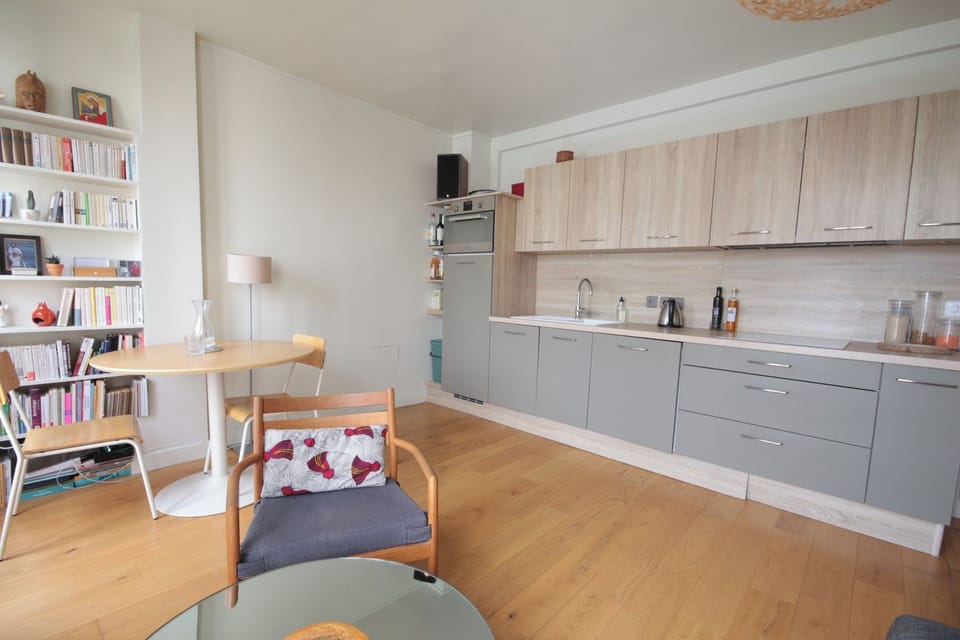 Cuisine équipée ouverte avec coin repas et bibliothèque, parquet clair — location Paris 19 près du Canal / Fully equipped open kitchen with dining nook and bookshelves, light hardwood floor — Paris 19 rental near the Canal