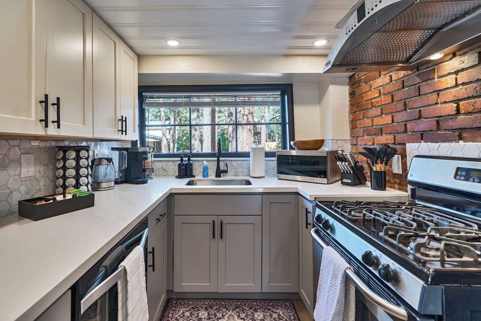 Sunlight pours through the picture window above the farmhouse sink, framed by exposed brick and gray shaker cabinetry. A well-stocked coffee station and gas range complete this bright, functional kitchen.
