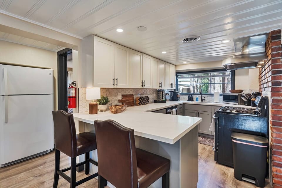 Leather barstools pull up to the crisp white peninsula, framed by full-size fridge storage and a sunny garden window. Exposed brick and a vintage range add character to this bright, well-equipped kitchen.