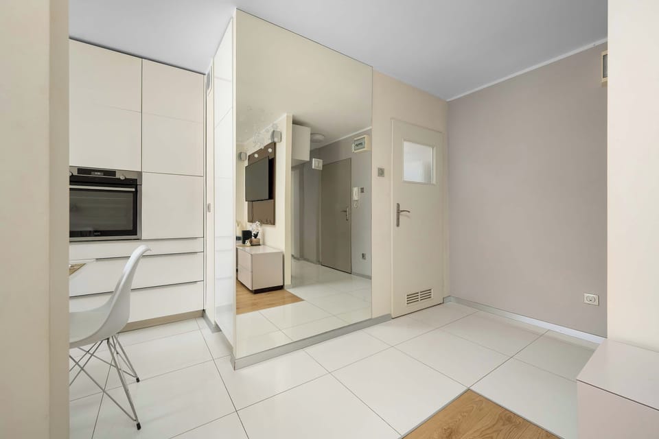 A bright open-plan room with a white TV cabinet and a television mounted on the wall. The dining area is visible farther back near the windows, and a doorway leads toward the kitchen or hallway.