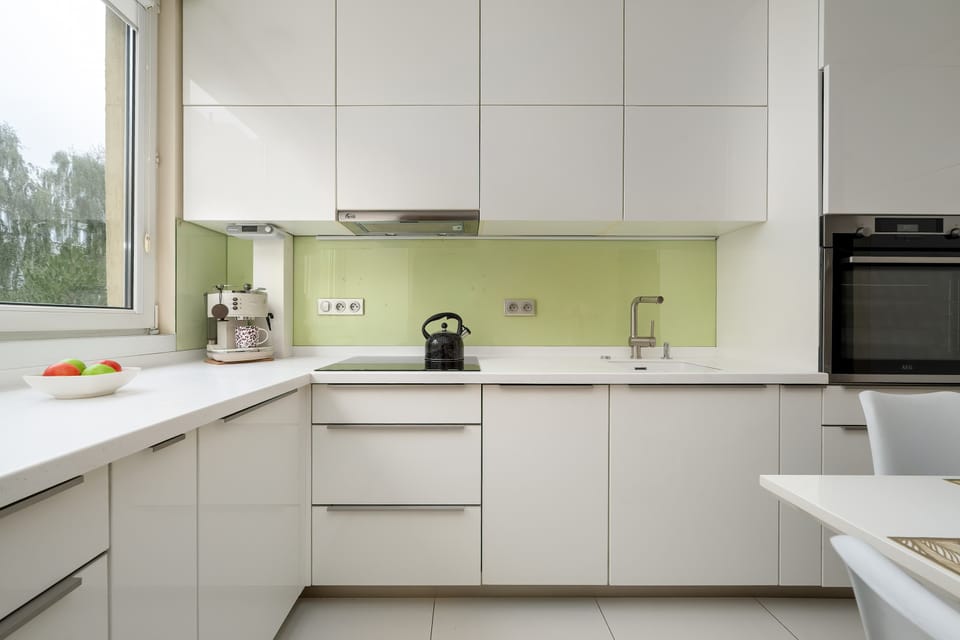 A modern white kitchen with sleek cabinets, a light countertop, and a pale green backsplash. A built-in oven and microwave are visible on the right, while a window provides a view of greenery outside.