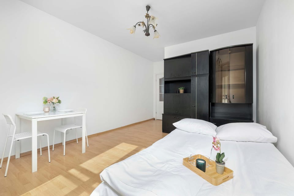 A bedroom view shows a dining table with two chairs and a sofa bed holding a breakfast tray. A large black wardrobe stands against the crisp white wall.