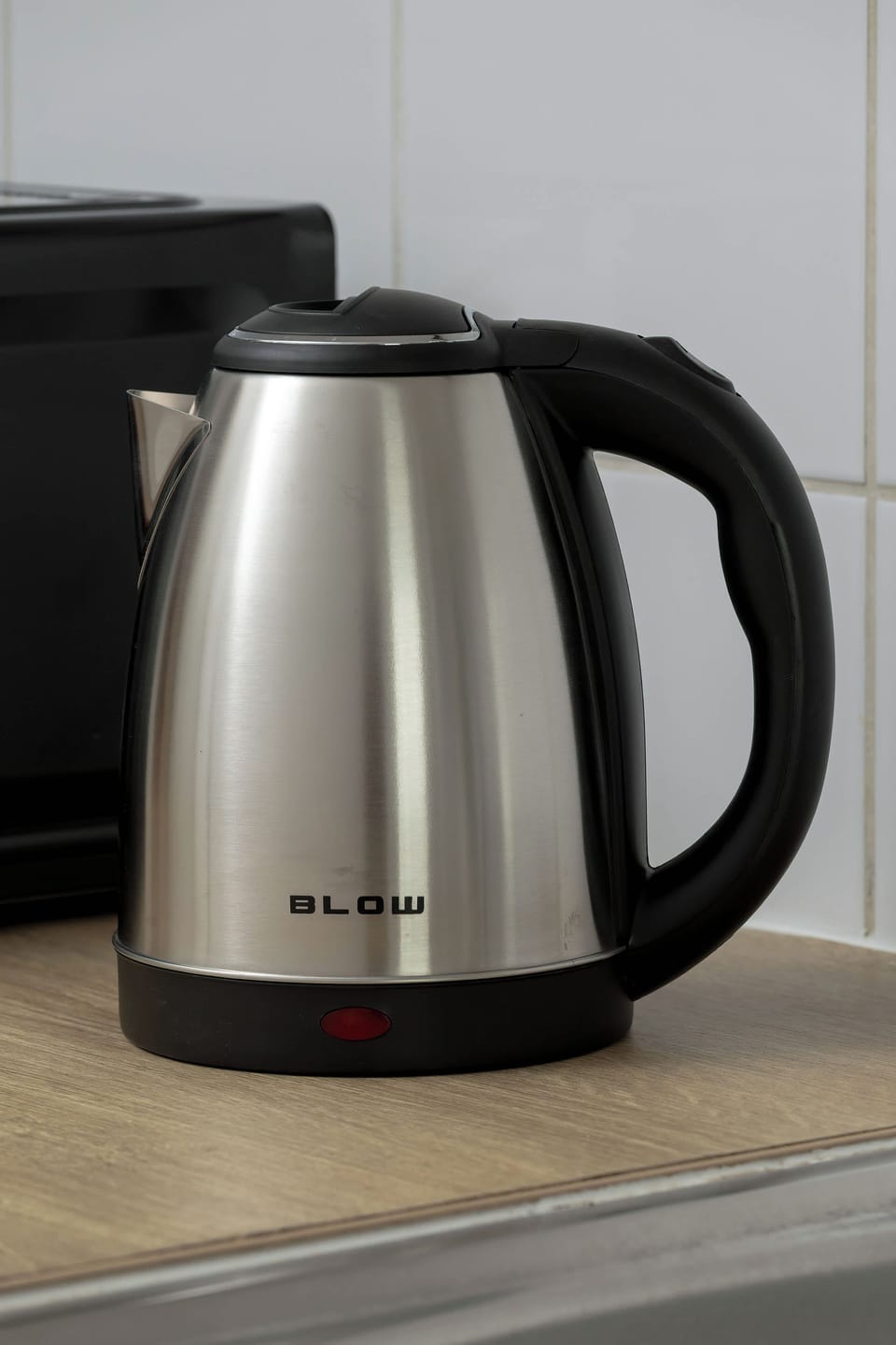 A close-up shot focuses on a stainless steel electric kettle resting on a kitchen countertop. Another black appliance is partially visible in the background.