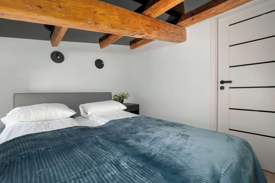 Another bedroom view centered on a large bed with white pillows and a blue-gray cover, beneath prominent exposed ceiling beams.