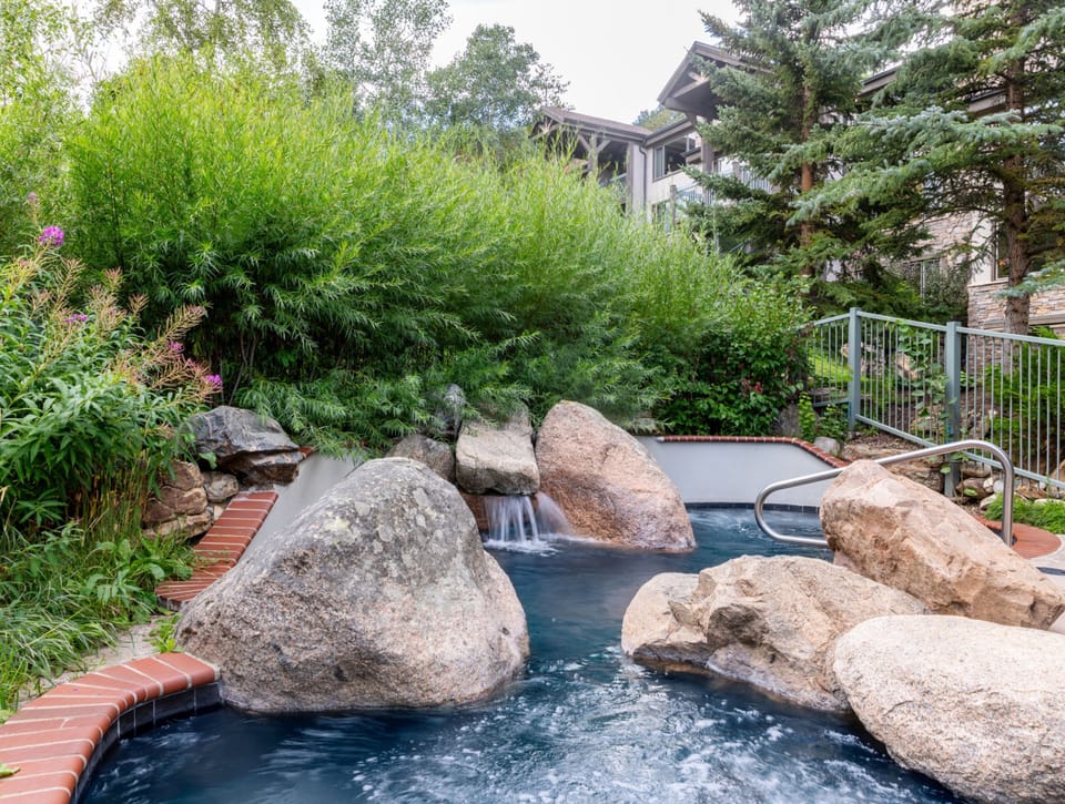 An outdoor hot tub surrounded by natural boulders and cascading waterfalls, nestled among lush greenery and evergreens, offers a serene mountain retreat.