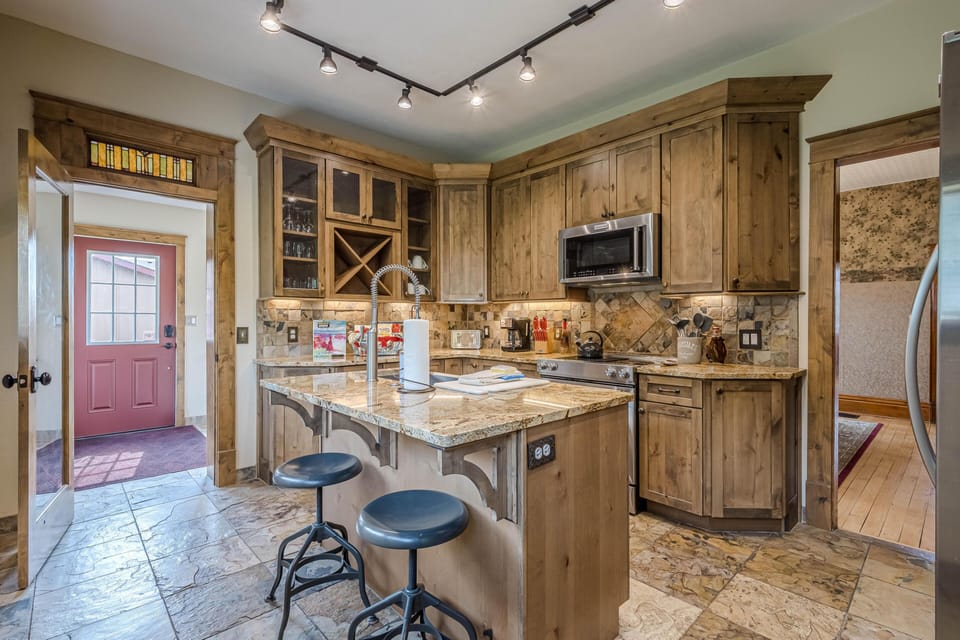 Natural wood cabinetry surrounds a granite-topped island with two metal stools and a high-arc faucet in the kitchen.