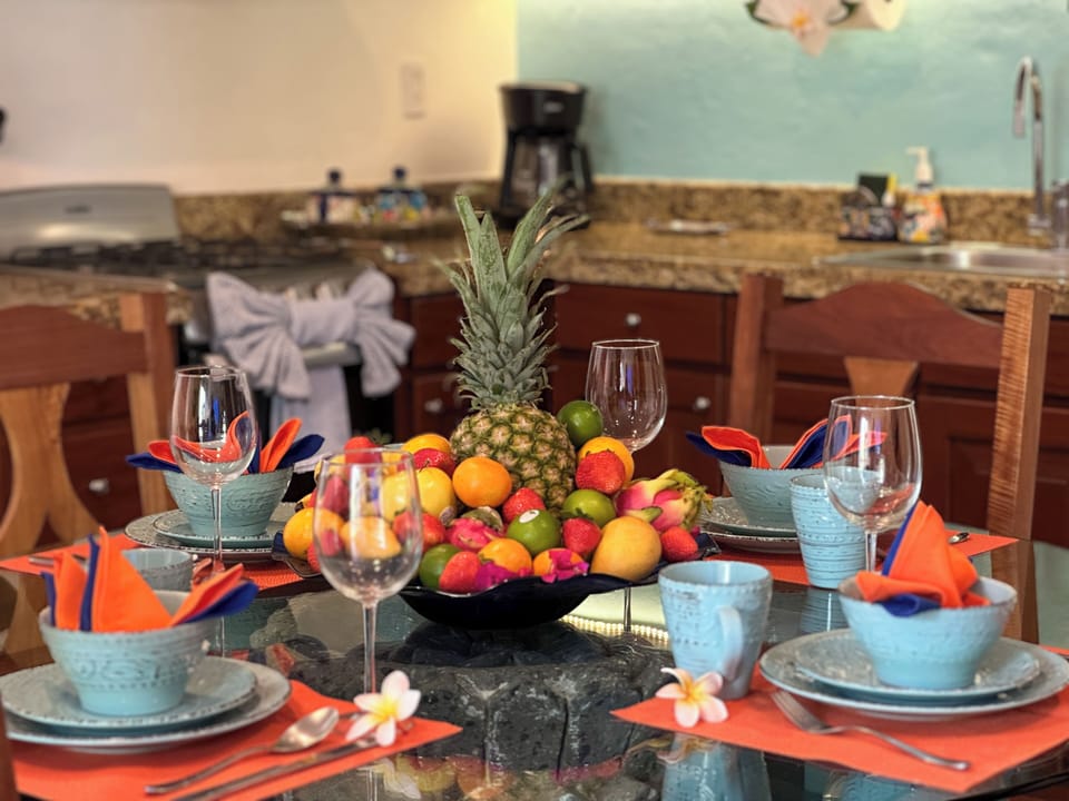 A2-10-2026 Dining Table - Dining area inside Abbey del Sol A2 Unit 10, a Puerto Morelos vacation rental, featuring colorful place settings, fresh fruit, wood cabinetry, and the kitchen in the background for an easy stay.
