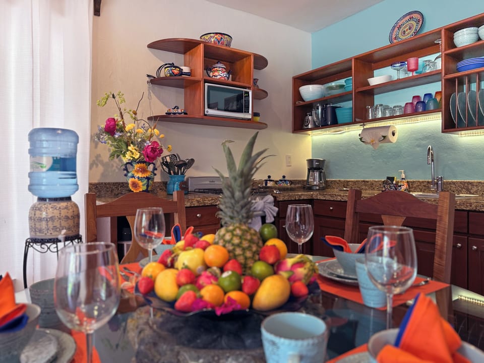 A2-10-2026 Kitchen and Dining Table - Kitchen and dining area inside Abbey del Sol A2 Unit 10, a Puerto Morelos vacation rental, featuring a dining table with fruit and place settings, wood cabinetry, open shelving, and a well-equipped kitchen for a comfortable stay.