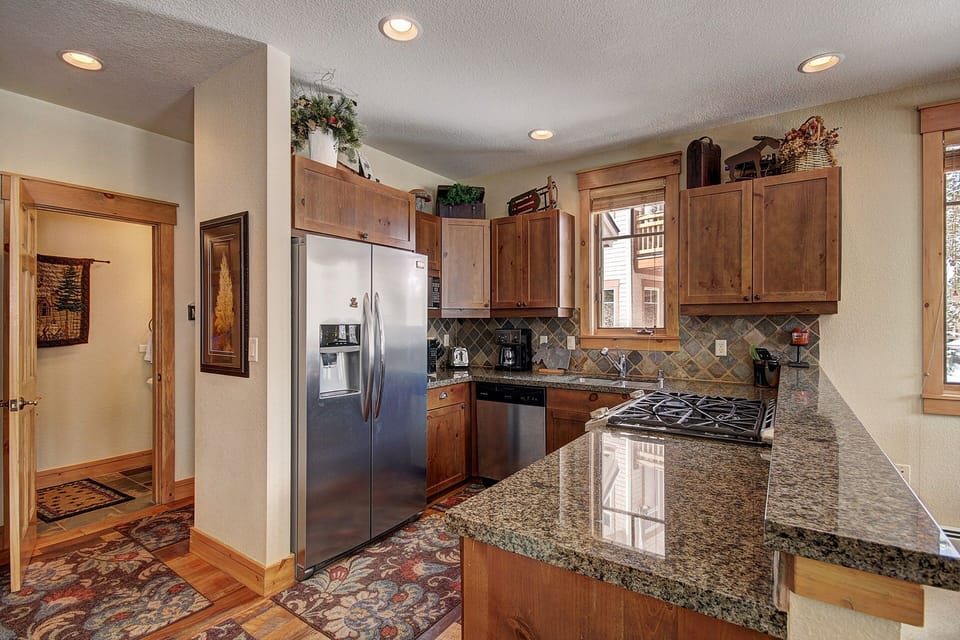 A modern kitchen with granite countertops, wooden cabinets, stainless steel appliances, and tiled backsplash. The space has a window, and there's a door leading to another room on the left.