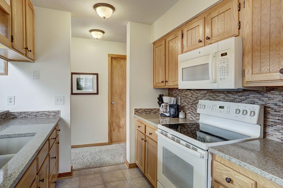 A kitchen with light wooden cabinets, a white electric stove, a microwave, and granite countertops. A hallway with carpeting and a framed picture is visible in the background.