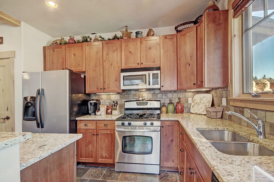 A kitchen with wooden cabinets, a stainless steel refrigerator, a gas stove, a microwave, and a coffee maker. Granite countertops and a window above the sink are also visible.