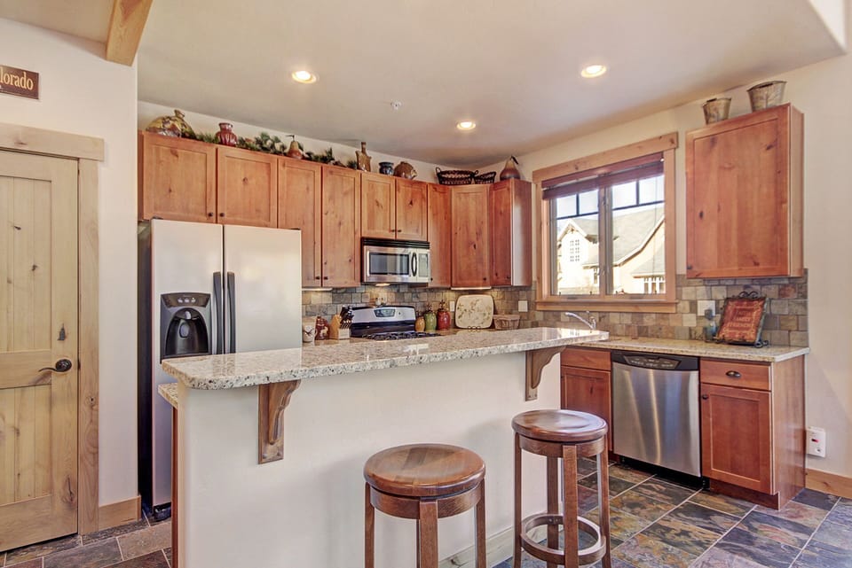 A cozy kitchen with wooden cabinets, granite countertops, a stainless steel refrigerator, oven, and dishwasher. The space includes a breakfast bar with two wooden stools and decorative items on top cabinets.