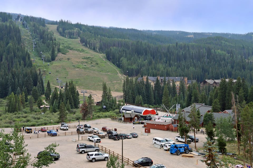 A mountainous area with skiing infrastructure, including ski lifts and a base lodge, surrounded by trees and a parking lot filled with vehicles at the bottom.