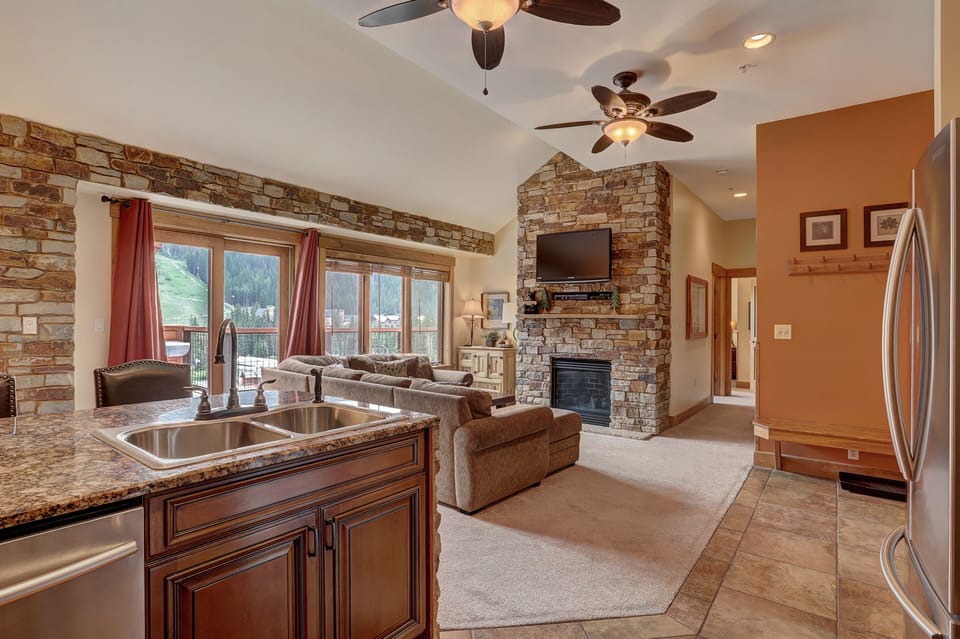 A cozy living room with stone walls and a fireplace, featuring a mounted TV, ceiling fans, and large windows. The room opens to a kitchen with a granite island and stainless steel appliances.