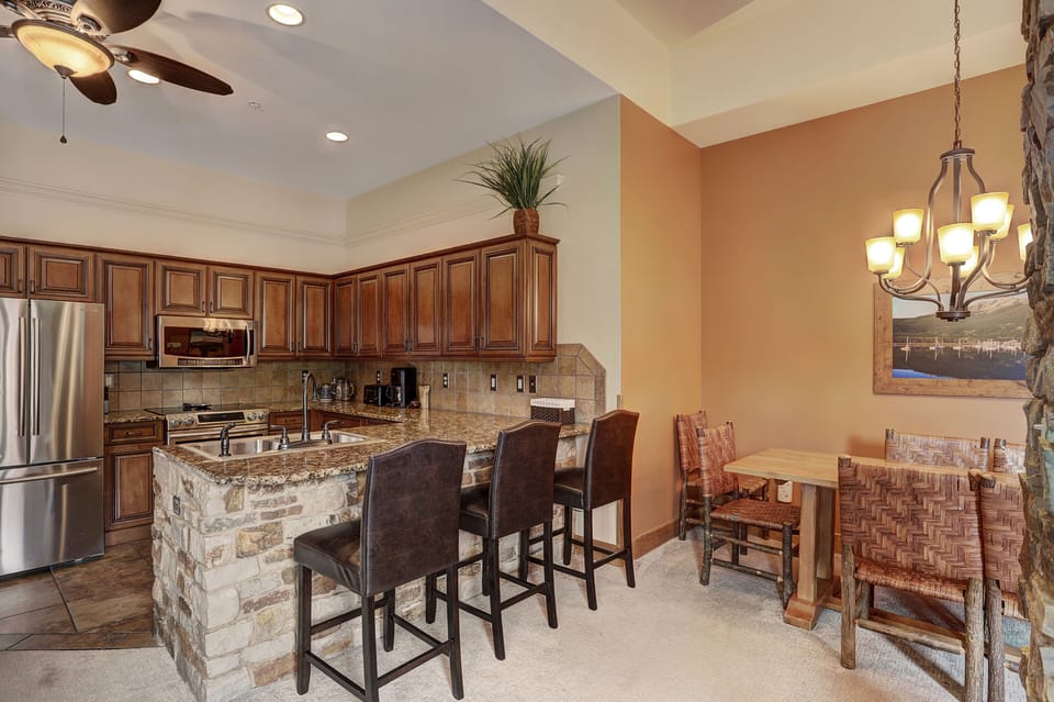 A kitchen and dining area with wooden cabinets, stainless steel appliances, an island with four bar stools, and a dining table with four chairs under a chandelier.