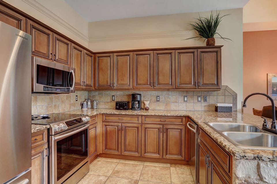 Modern kitchen with wooden cabinets, stainless steel appliances, a double sink with granite countertops, and a tiled backsplash. A potted plant is placed on top of the cabinets.