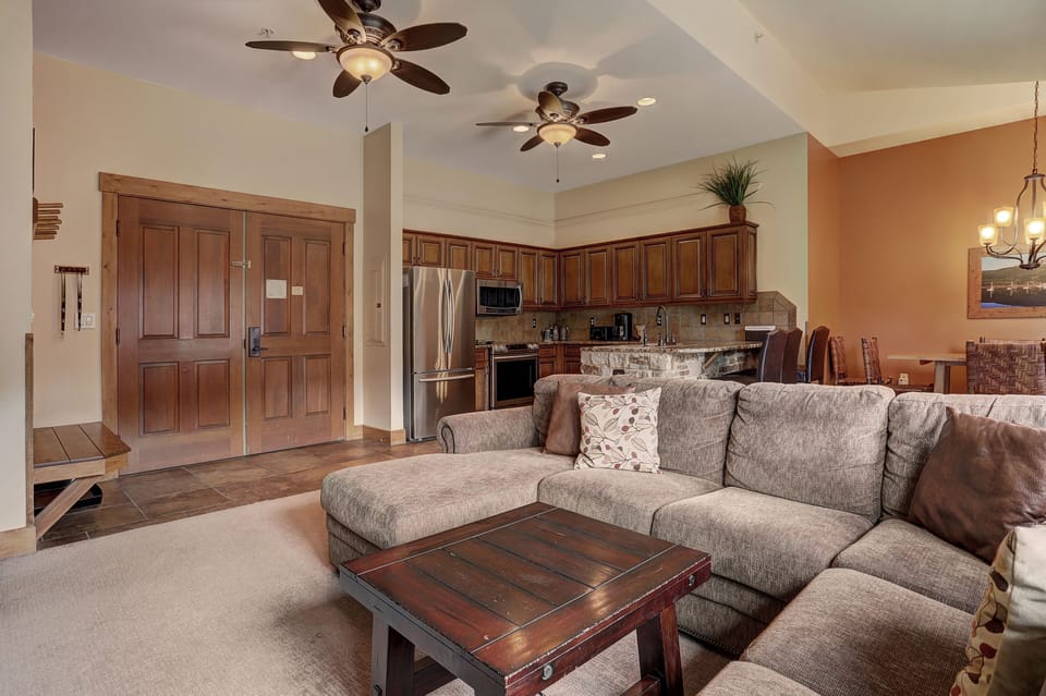 Open-concept living room with a large sectional sofa, wooden coffee table, and ceiling fans. The room extends to a kitchen with wooden cabinetry and stainless steel appliances.