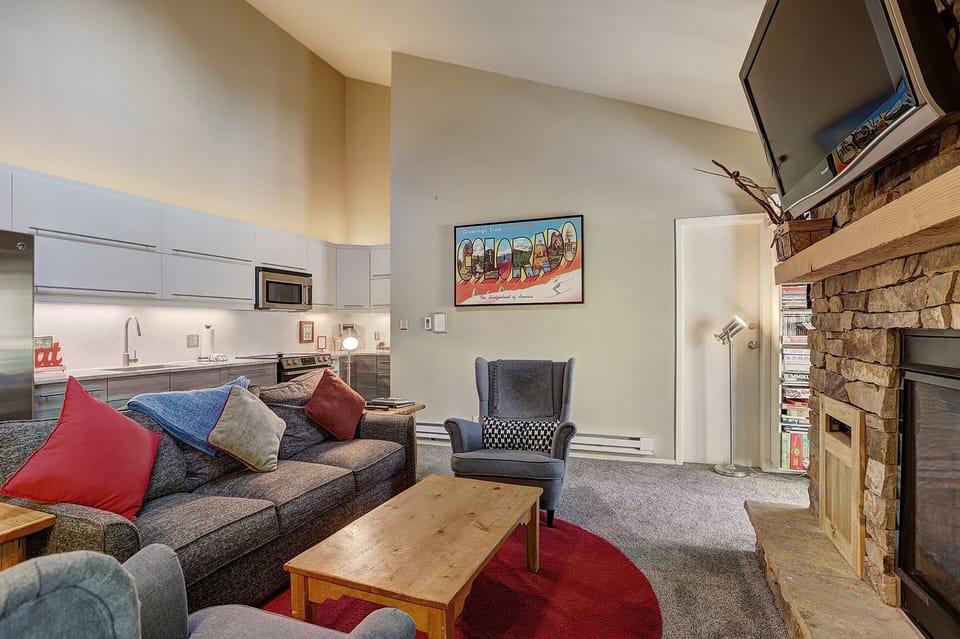 A cozy living room with a gray sofa, colorful throw pillows, a wooden coffee table, and an armchair. The kitchen is visible in the background, and there's a fireplace with a TV mounted above it.