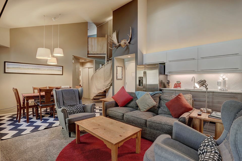 A modern living room and dining area with a gray sofa, colorful cushions, armchairs, wooden coffee table, dining table with chairs, and spiral staircase leading to a loft. A mounted deer head decorates the wall.