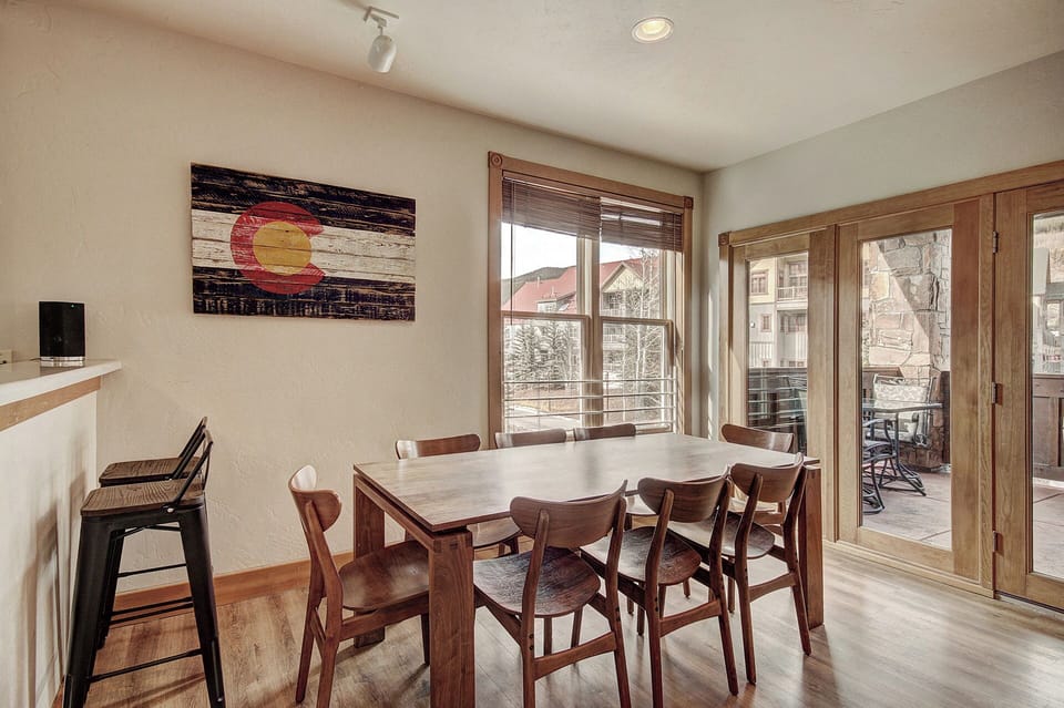 A dining area with a wooden table and six chairs, a wall-mounted Colorado flag, large windows, and a door opening to a patio with outdoor seating.